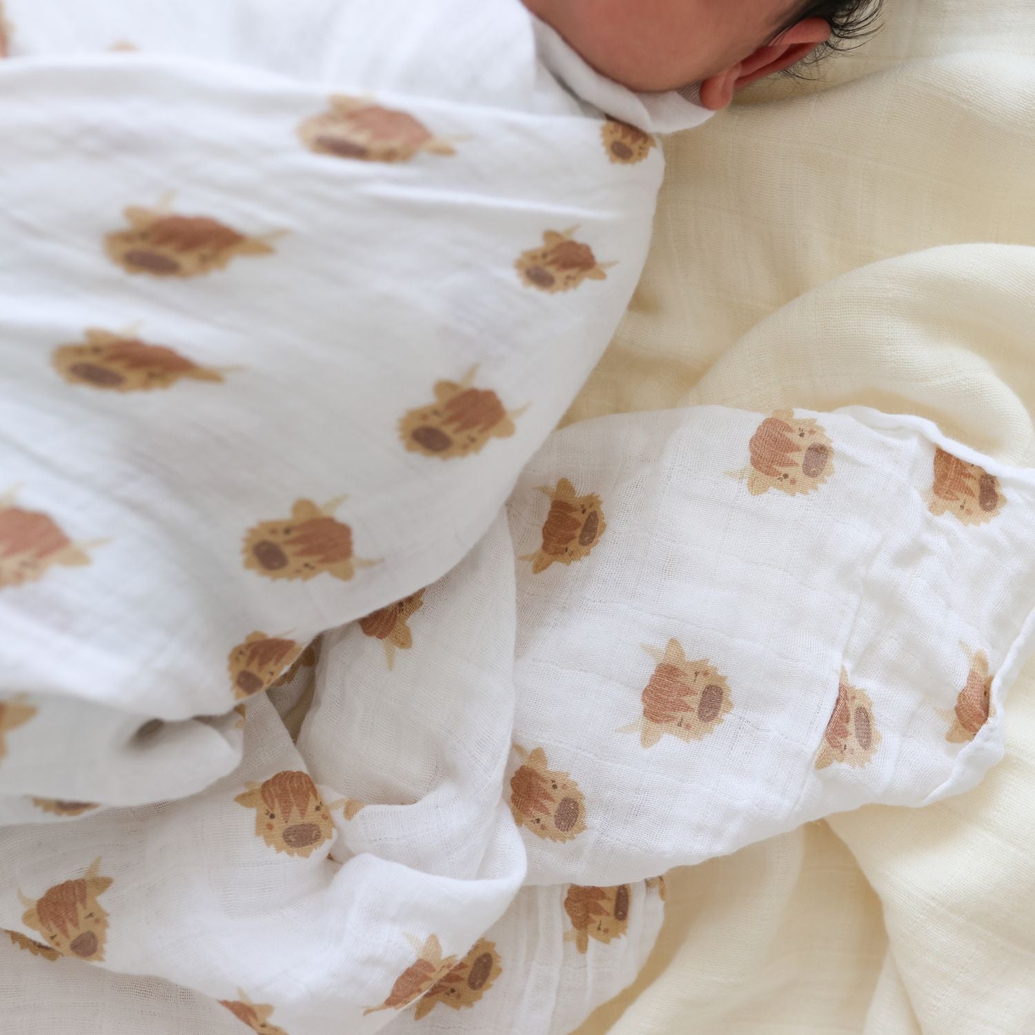 Baby swaddled in a white blanket with highland cow animal prints on a soft beige surface.