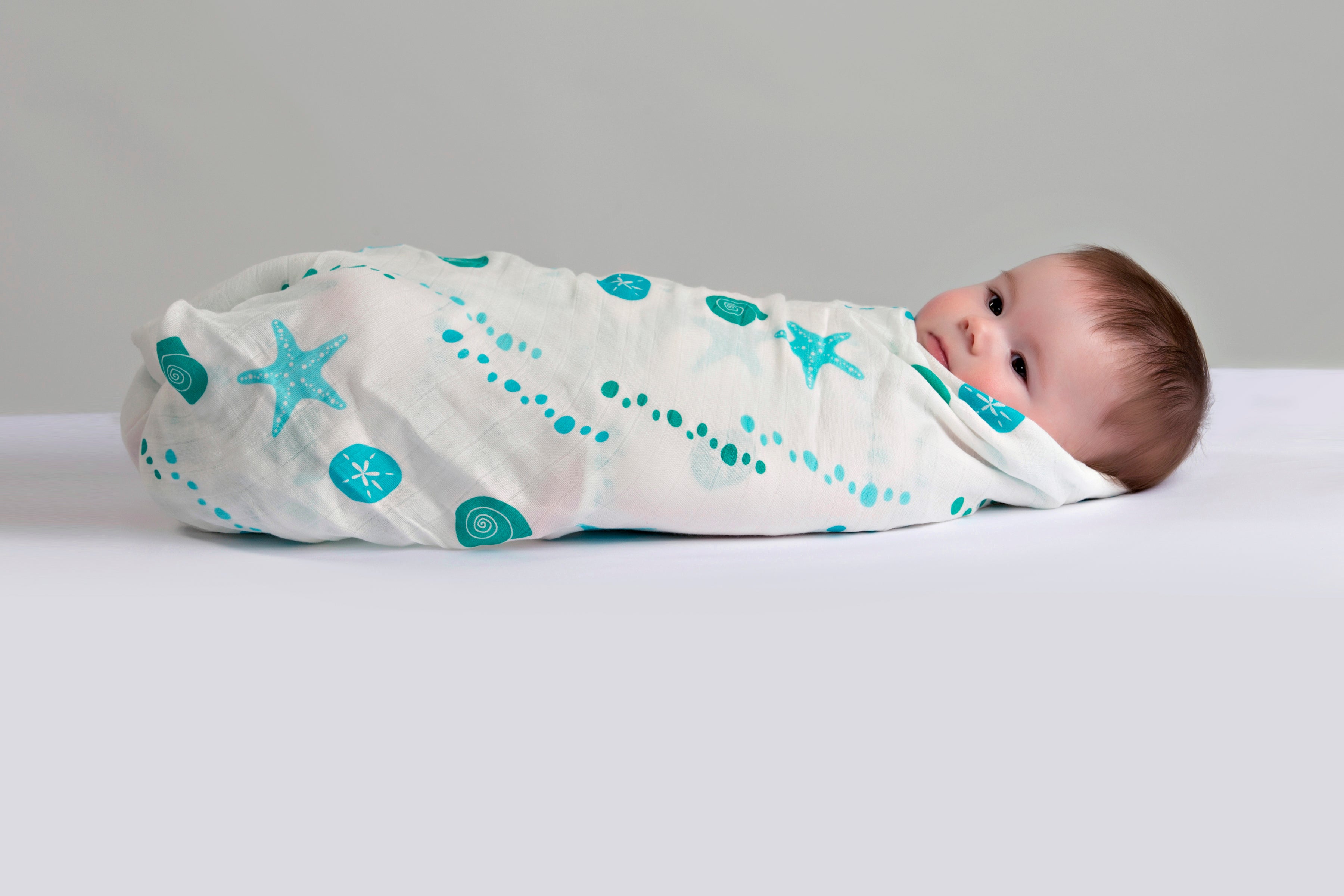 Baby swaddled in a white muslin blanket with turquoise blue sea shells and starfish ocean pattern